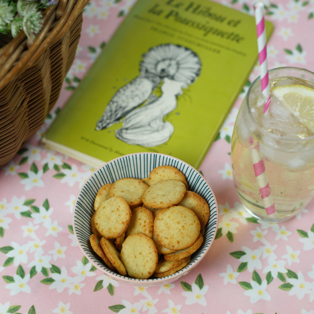 A bowl of original cheese sables on a table next to a  summer drink and a book.
